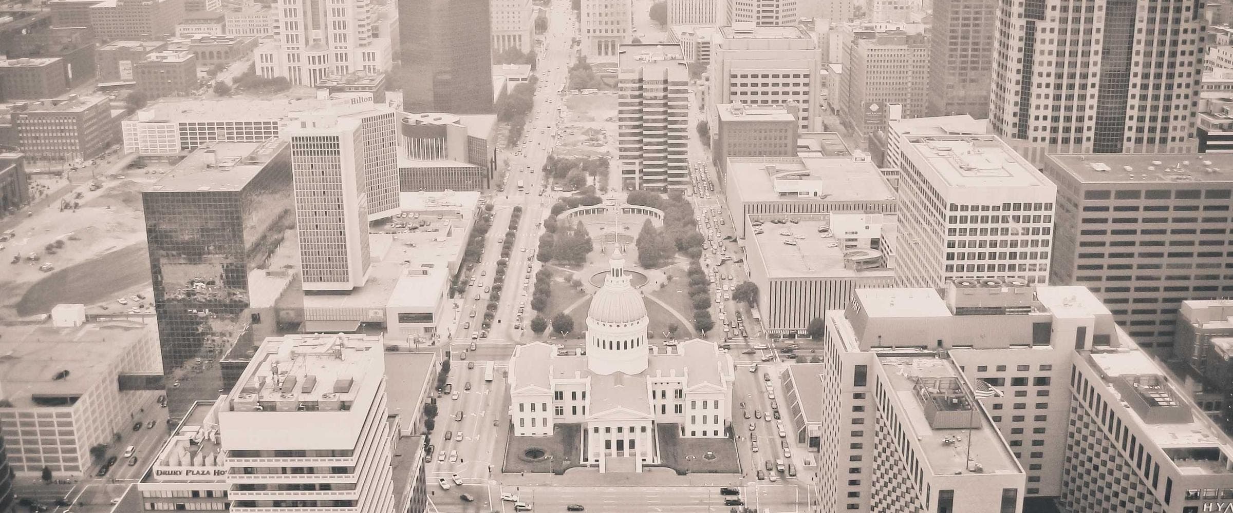 a downtown from high above in warm sepia, a domed courthouse at the head of a long boulevard with traffic threading between the towers
