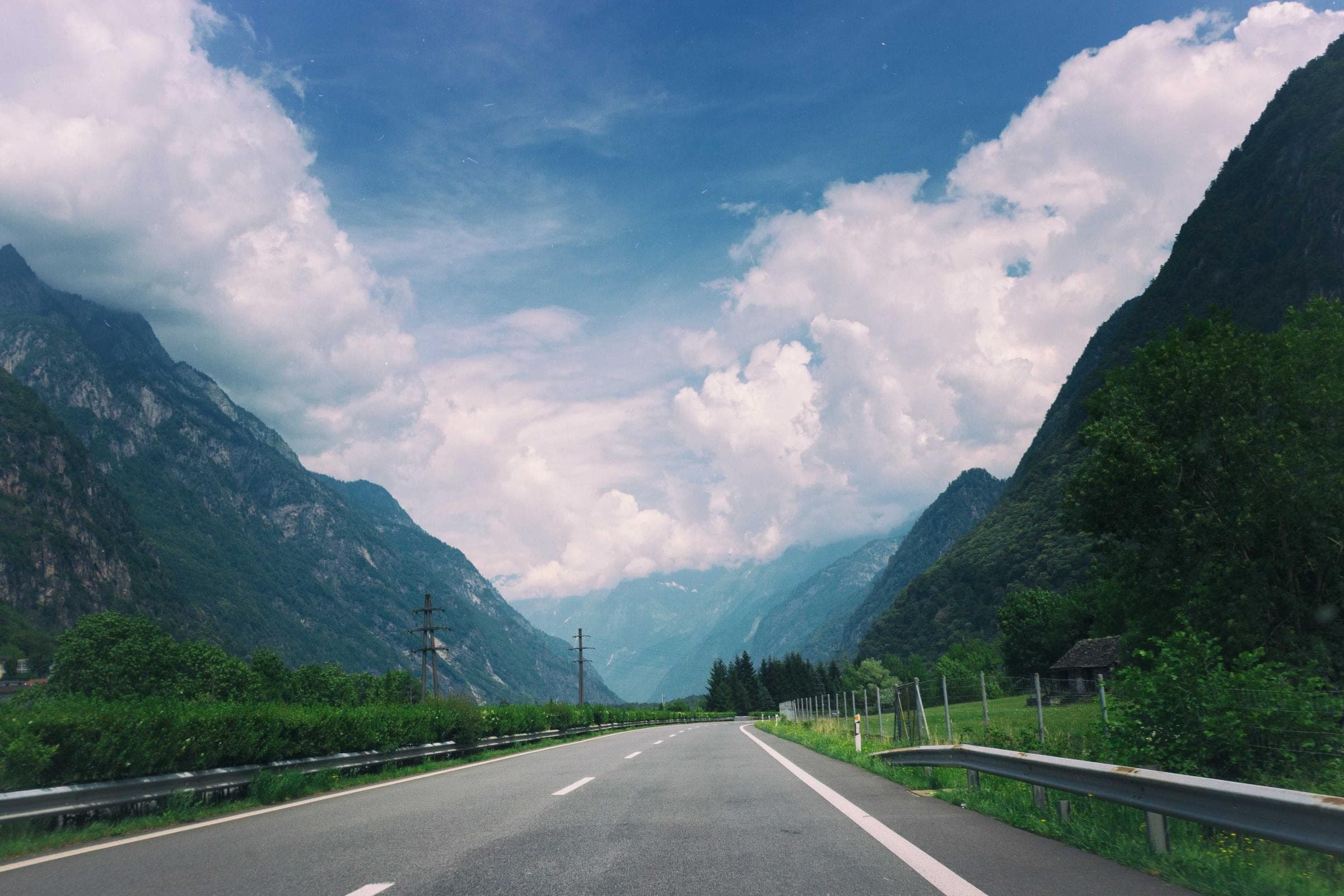 an empty two-lane road running toward a steep green valley, tall cumulus stacked above the ridgelines on either side