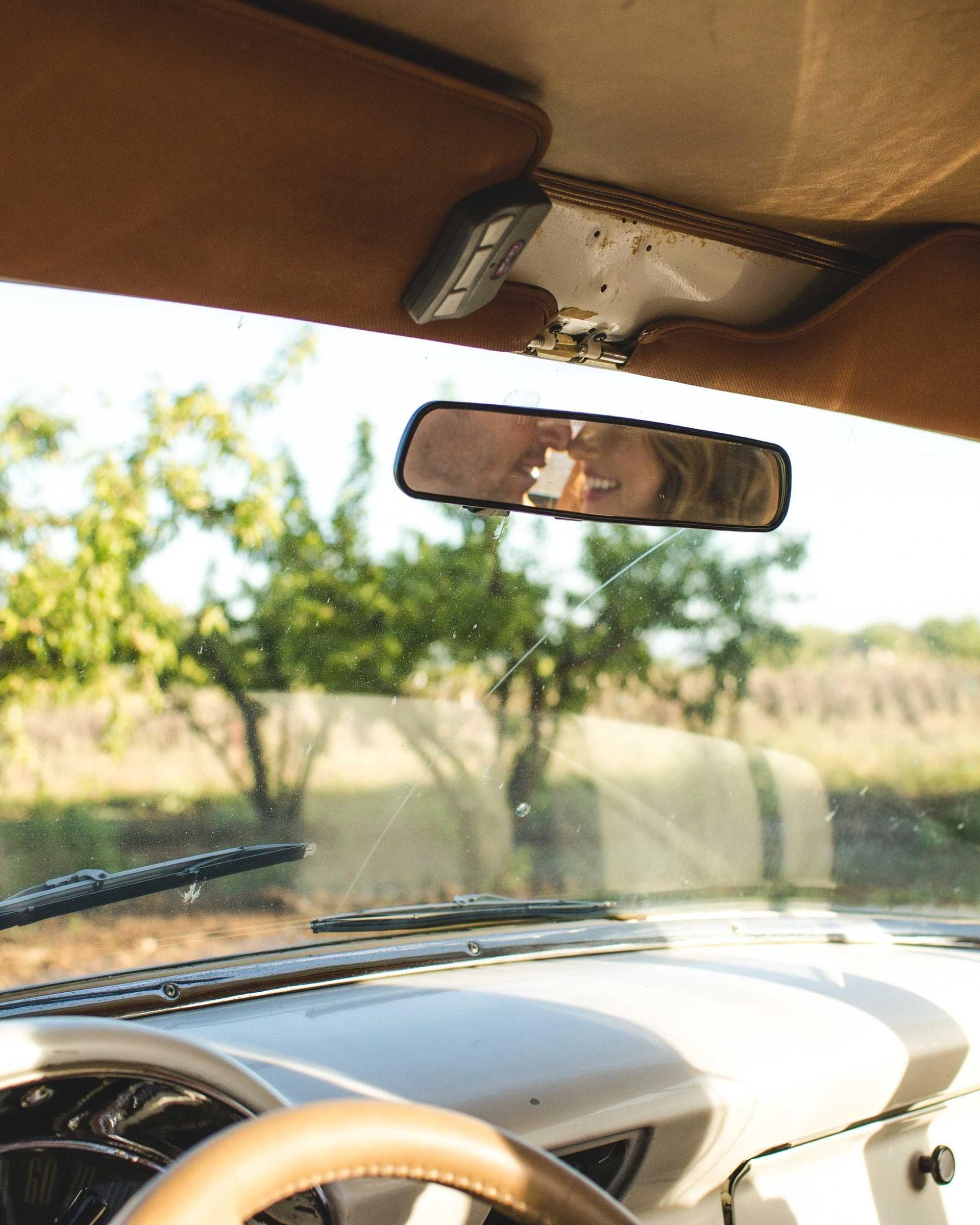 the inside of an old convertible in low sun, the rear-view mirror holding two people laughing, an orchard beyond the windshield