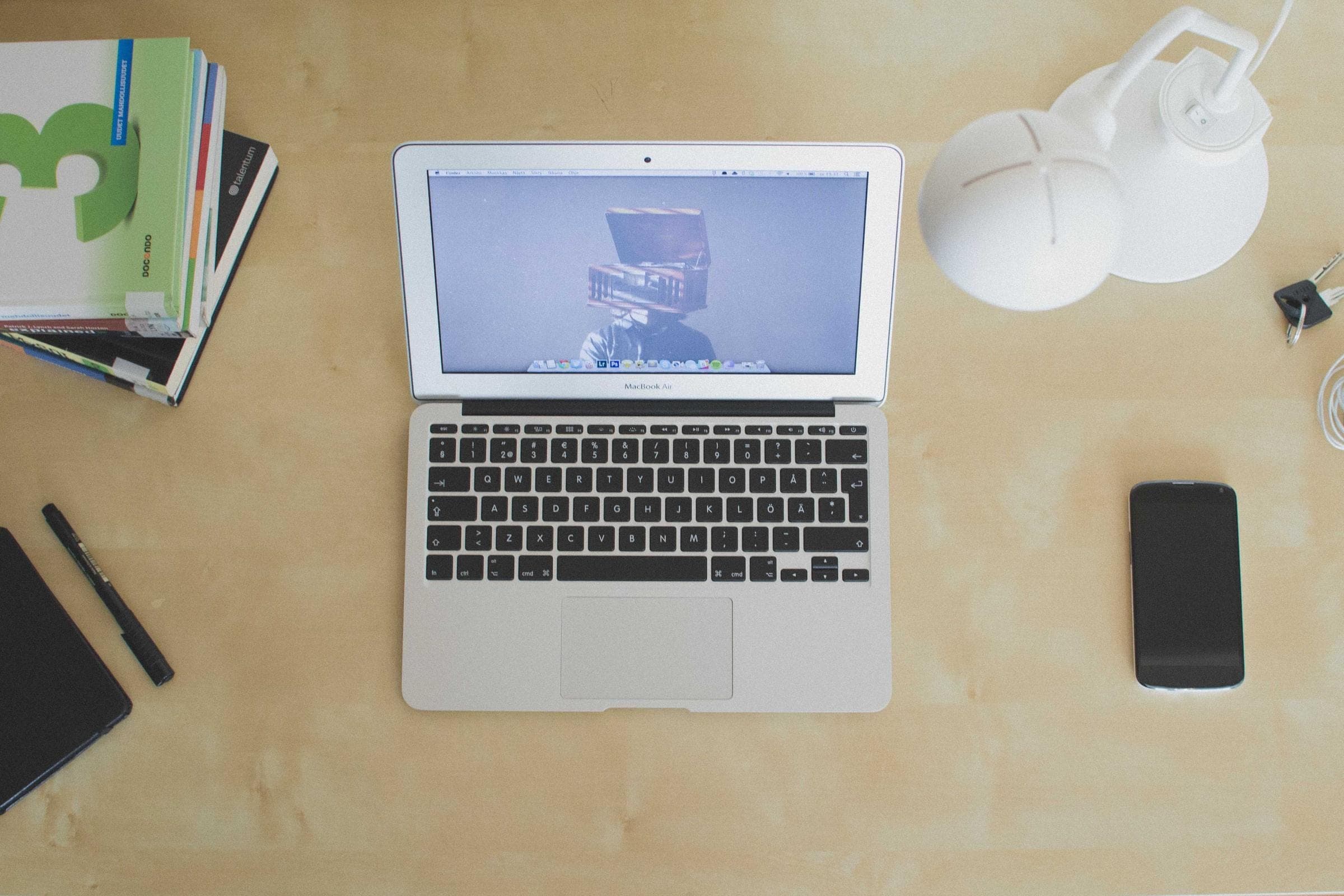 a desk from directly overhead, an open laptop centered on pale wood with a stack of books, a lamp, a phone and keys around it