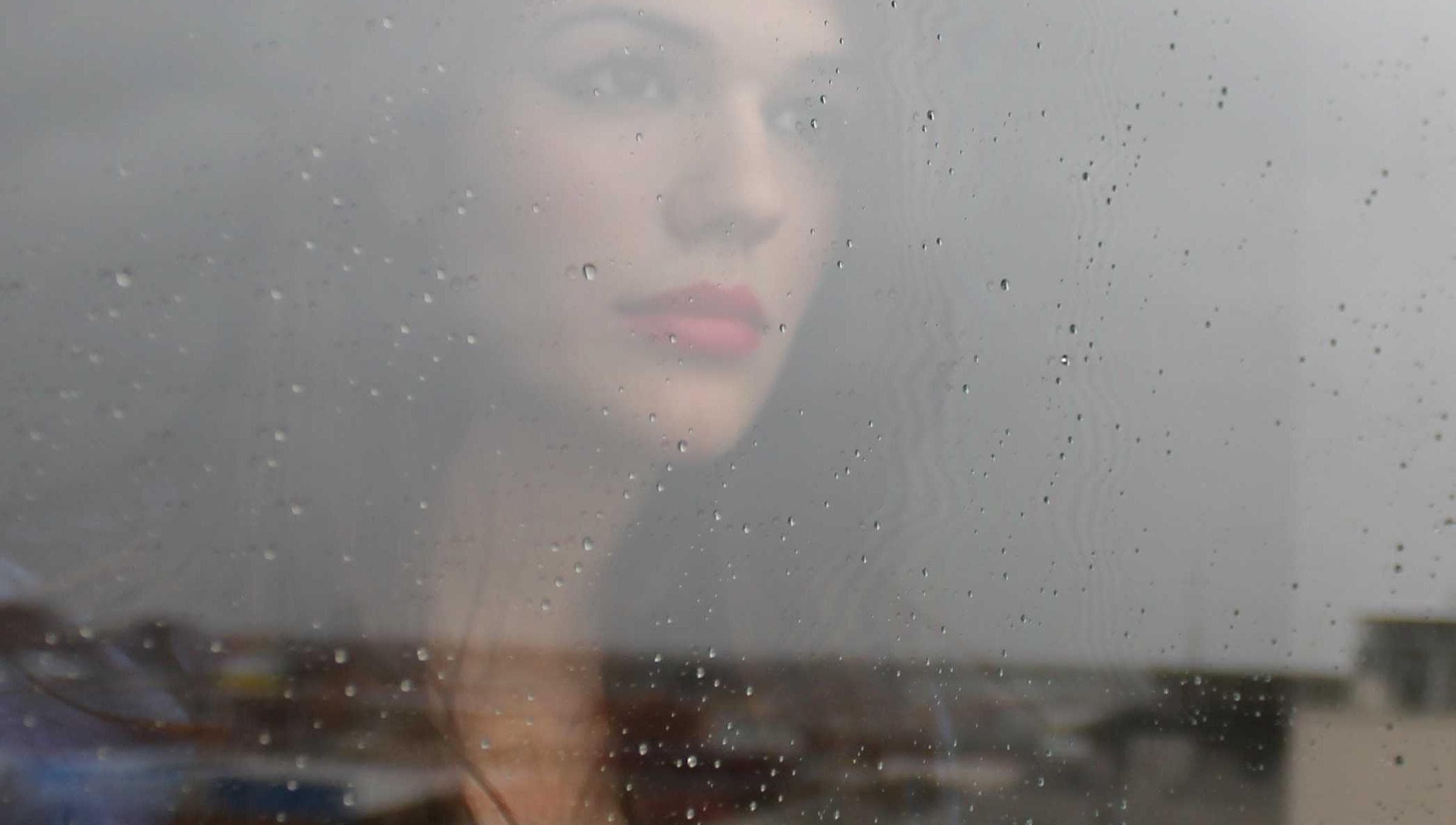 a face behind a rain-flecked pane, softened by the glass, grey sky and rooftops along the bottom of the frame