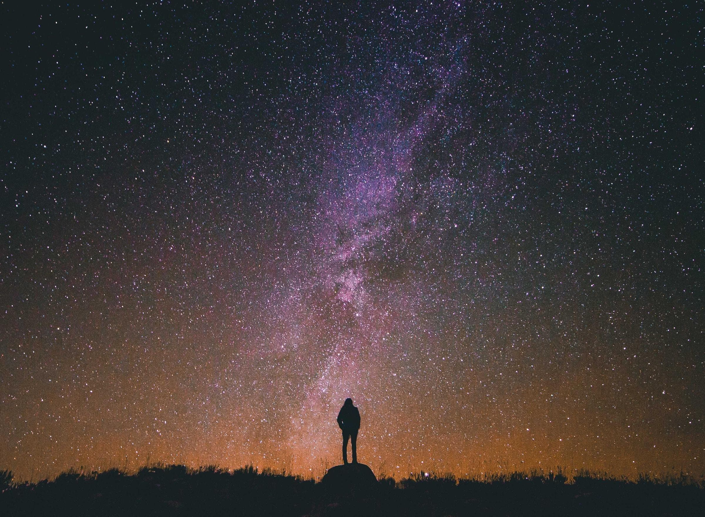 the milky way standing over a dark horizon, one figure on a boulder silhouetted against the glow at the skyline